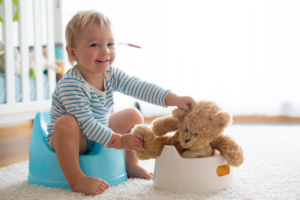 child sitting on potty chair. He holds a stuffed animal bear that is also sitting on its own potty chair. Shows that making going potty fun and relaxed helps children learn to be comfortable on potty.